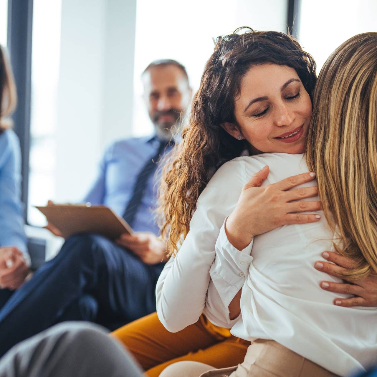 A woman warmly hugging another during a group therapy session.
